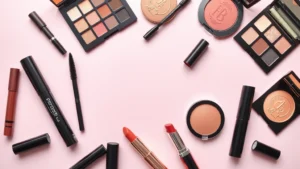 Flat lay arrangement of colorful affordable makeup products including mascaras, eyeshadow palettes, lipsticks, and blush compacts on a soft pink background with natural lighting, no text or labels visible, professional beauty photography style