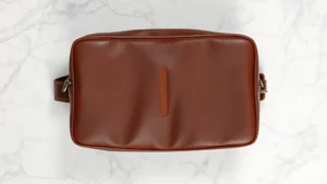 High-quality full-grain brown leather Dopp kit photographed from above on white marble surface, showing rich patina and texture detail, soft natural lighting highlighting leather grain