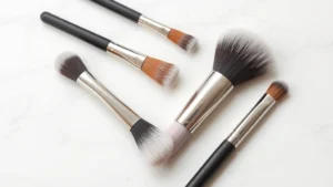 Close-up of five essential makeup brushes arranged flat lay on white marble surface, showing natural hair textures and metal ferrules, professional studio lighting