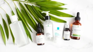 Flat lay of beginner skincare essentials: cleanser, toner, moisturizer, sunscreen, and serum bottles arranged on white marble surface with green plant leaves and soft natural lighting